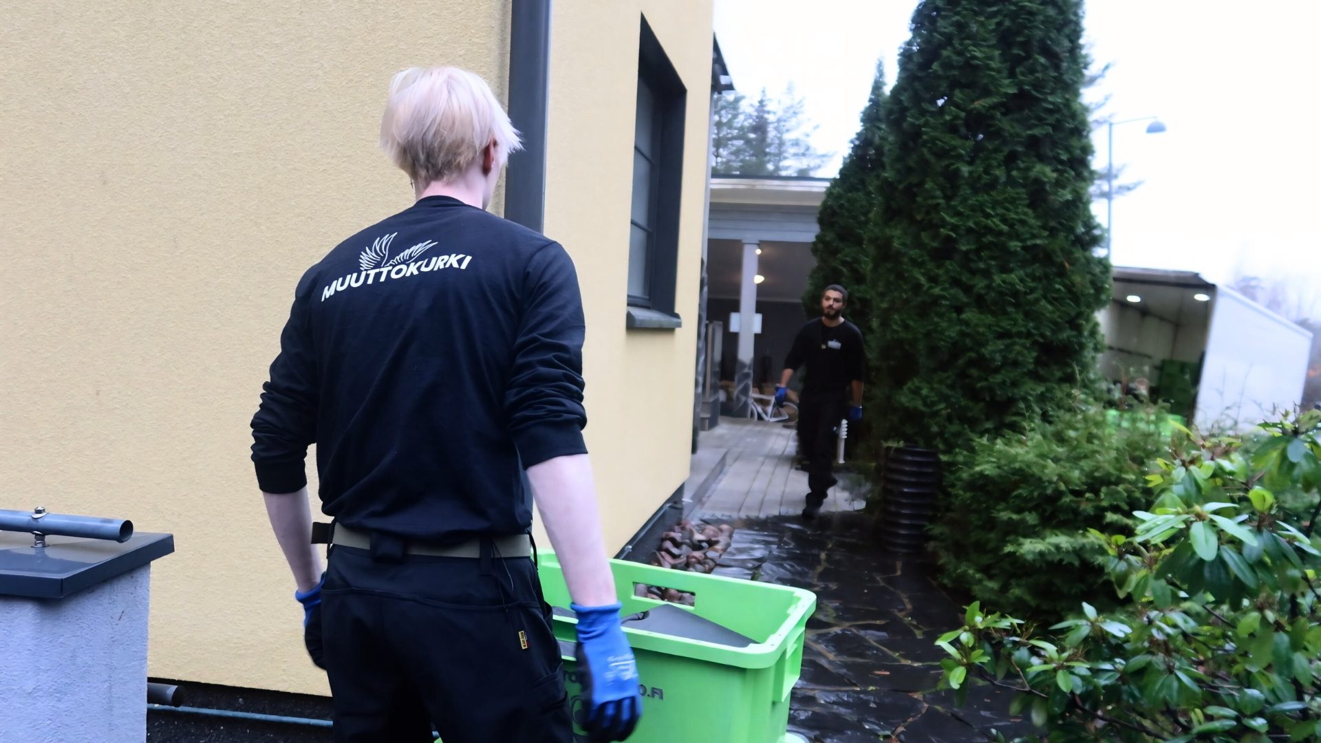 Muuttokurjen työntekijä kantaa vihreitä muovilaatikoita talon pihalla.